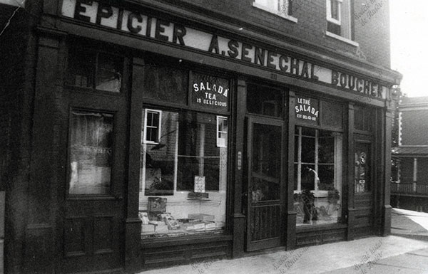 Épicerie A. Sénéchal. Les fenêtes de la boulangerie Lacombe, en face, refletent dans la vitrine de la bâtisse.